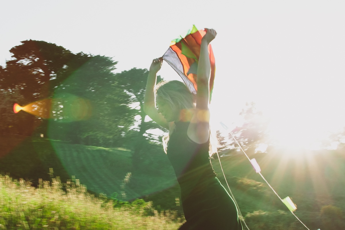 Musician Anya Anastasia dancing in a sunny outdoor field holding a colorful fabric overhead, backlit by golden sunlight with lens flare.